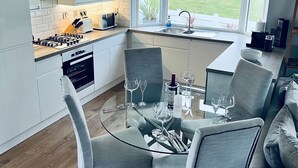 Een moderne keuken met witte kastjes, een gasfornuis, en een eethoek met een glazen tafel en grijze stoelen.