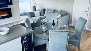 Een moderne eethoek met een glazen tafel, grijze bekleden stoelen en een televisie waarop een haardbeeld wordt vertoond.