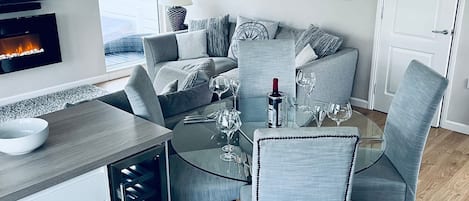 Un espace repas moderne avec une table en verre, des chaises à revêtement gris et un téléviseur diffusant une image de cheminée.