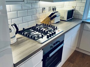 A modern kitchen with white cabinets, a gas stove, a microwave, and a toaster. - The Warren O7 (Pwllheli)