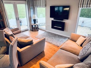A modern living room with a sofa set, a flat-screen TV mounted on the wall, and a view of the outdoors through large windows. - The Warren O7 (Pwllheli)