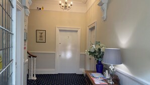Um corredor com uma porta identificada com o número 2, um candelabro, um vaso azul com flores e um abajur sobre uma mesa de madeira.