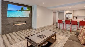 A modern living room with a fireplace, a flat-screen TV, a dining area with red chairs, and a kitchen in the background.