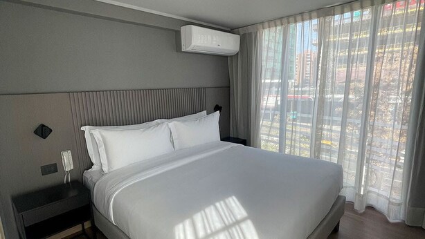 Ein modernes Hotelzimmer mit einem großen Bett, einem Nachttisch, einer Klimaanlage und einem fenster mit Vorhängen.