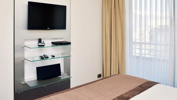 A hotel room with a flat-screen TV mounted on a wall, a glass shelf with electronic devices, a bed with a brown headboard, and a window with sheer curtains. - Manquehue Retreat EXC Stylish Home in Las Condes (Santiago)