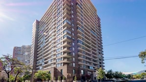 A high-rise residential building with multiple balconies, a clear sky, and surrounding greenery.