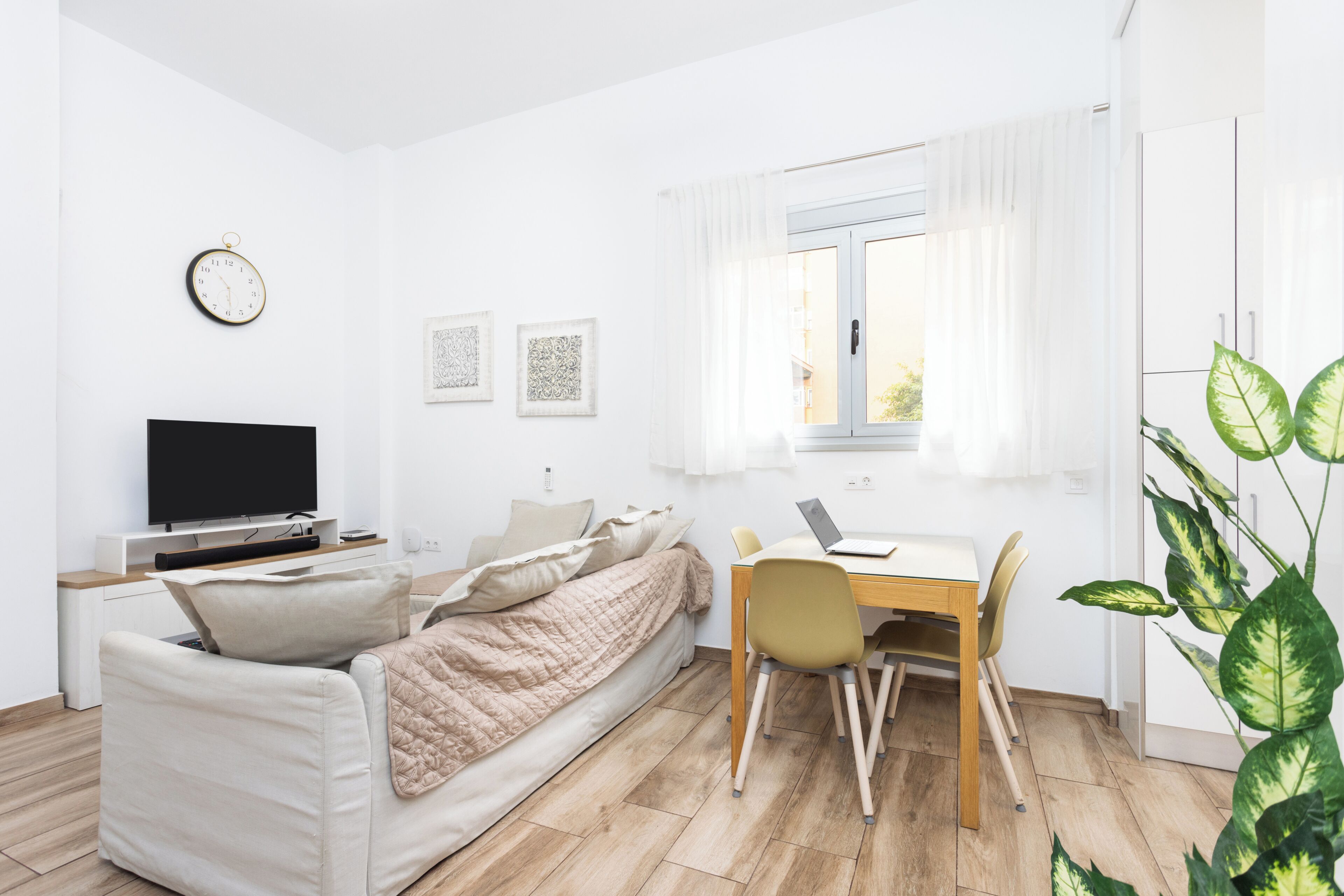 Una habitación moderna y luminosa con una cama, un televisor, un escritorio con una computadora portátil y una ventana con cortinas.