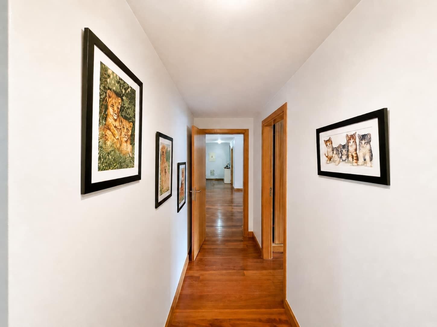 A hallway with framed pictures on the walls, wooden flooring, and a door leading to another room.