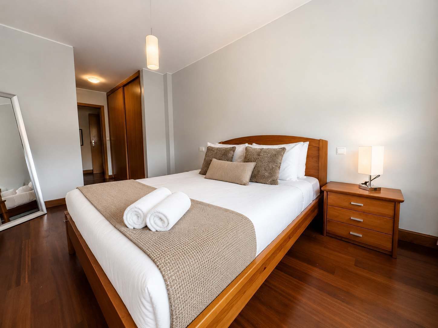 Un lit bien fait, avec une tête de lit en bois, une table de chevet avec une lampe et un grand miroir au mur.