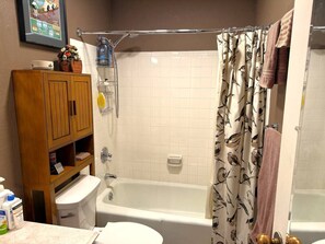 A bathroom with a bathtub, shower curtain, wooden cabinet, and a shelf with toiletries. - EagleVail Golf Course Cozy Retreat | Ground Floor (Avon)