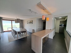 A modern kitchen with a central island, a dining area with a table and chairs, and a view through a glass door to an outdoor area. - Family Apartment in Groenendijk (Koksijde)