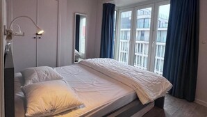 Ein Schlafzimmer mit Bett, Nachttischlampe, einem Fenster mit Vorhängen und Blick auf Gebäude.