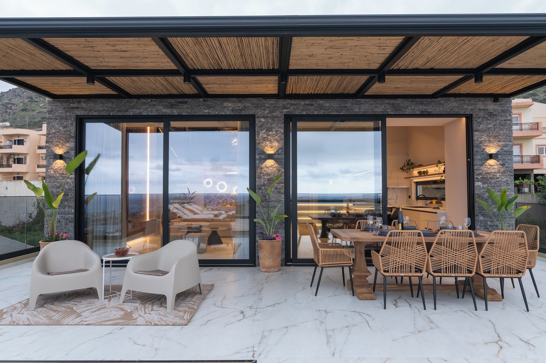 A modern outdoor dining area with a view of the sea, a stone wall, and a glass door leading to a kitchen.