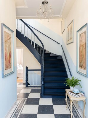 A grand staircase with a black and white checkered floor, a chandelier, and framed artwork on the walls.