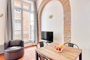 A room with a brick wall, a television on a stand, a wooden table with fruit, a leather chair, and a window with curtains. - Duplex T2 (Unit 5B) overlooking Place du Capitole (Toulouse)