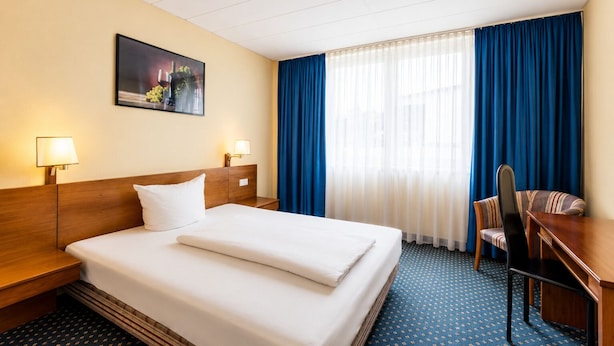 Een hotelkamer met een bed, twee nachtkastjes, een bureau met stoel, een raam met blauwe gordijnen en een schilderij aan de muur.