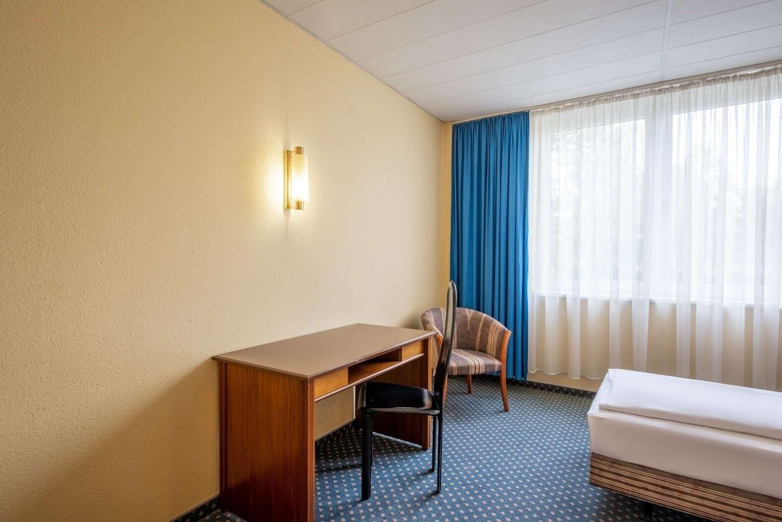 Una habitación de hotel con un escritorio de madera, una silla, una cama y una ventana con cortinas azules.