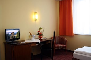 Habitación de hotel con una cama, un escritorio con televisor, una silla, un jarrón con flores y una lámpara fijada a la pared.