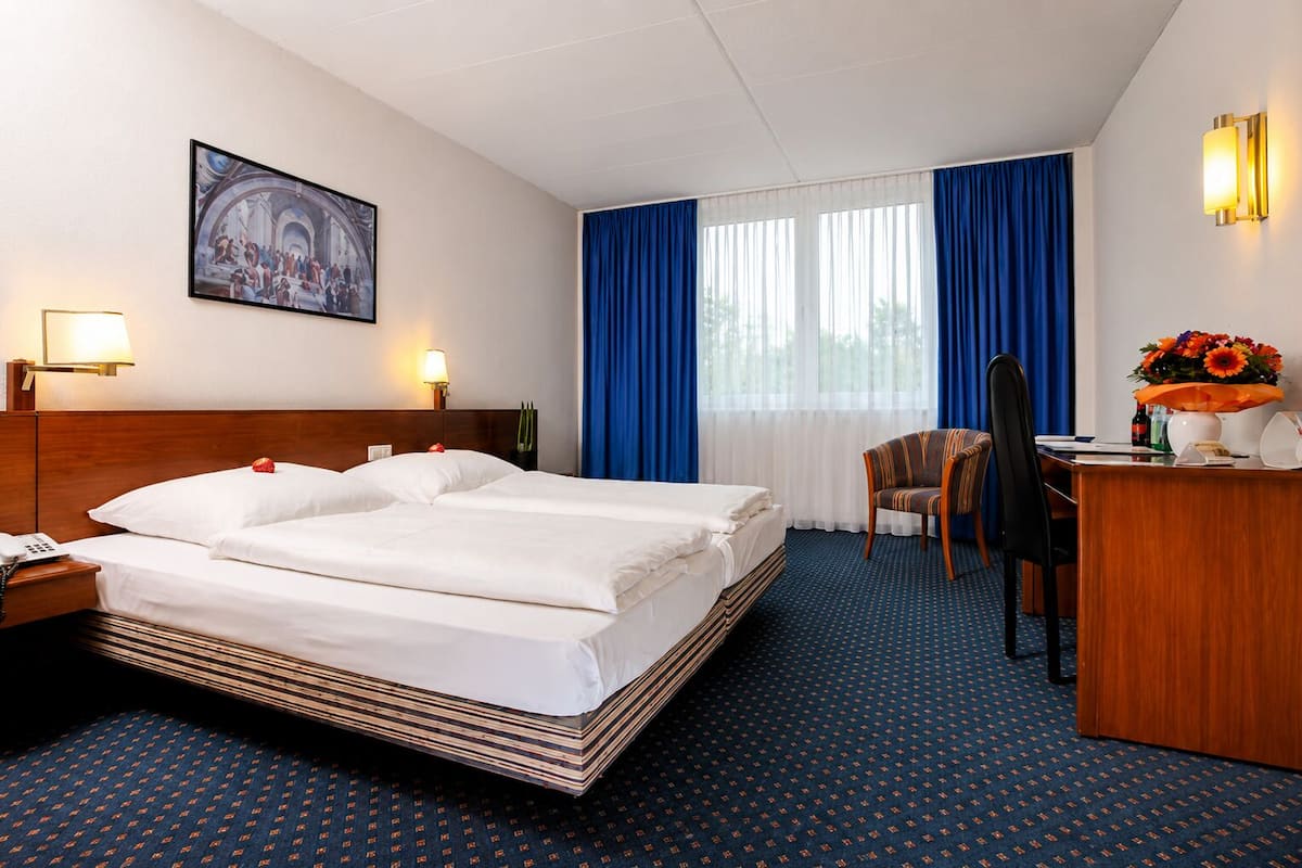 Ein Hotelzimmer mit einem Doppelbett, einem Schreibtisch mit Telefon, einem Stuhl, einer Schreibtischlampe, einem Bild an der Wand und einem Fenster mit blauen Vorhängen.