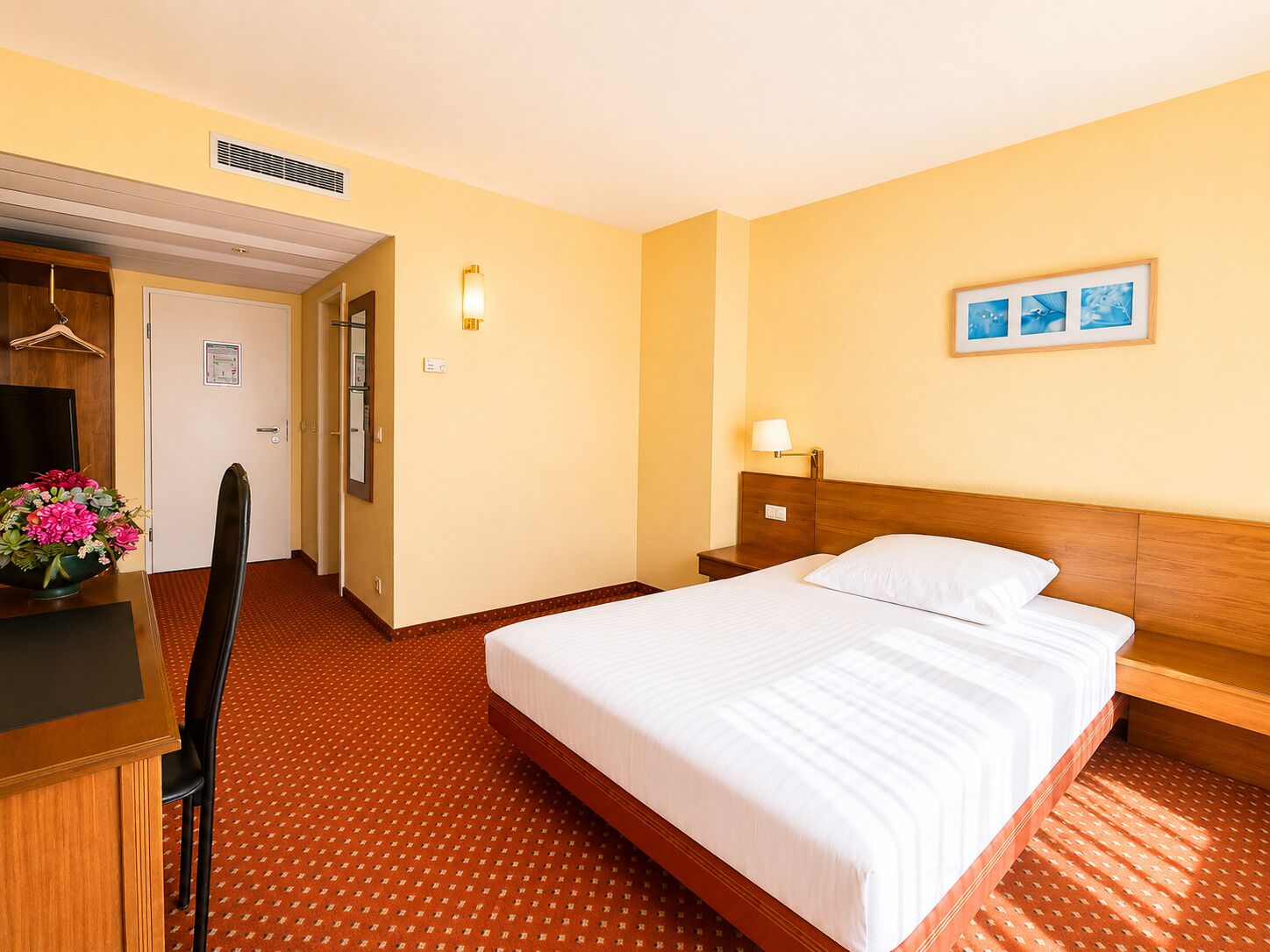 Hotellihuone, jossa on sänky, työpöytä tuolilla, yöpöytä, seinään kiinnitetty valaisin ja seinällä oleva kehystetty kuva.