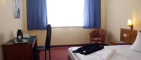 A hotel room with a bed, a desk with a laptop, a chair, a small table with a lamp, and a window with curtains.