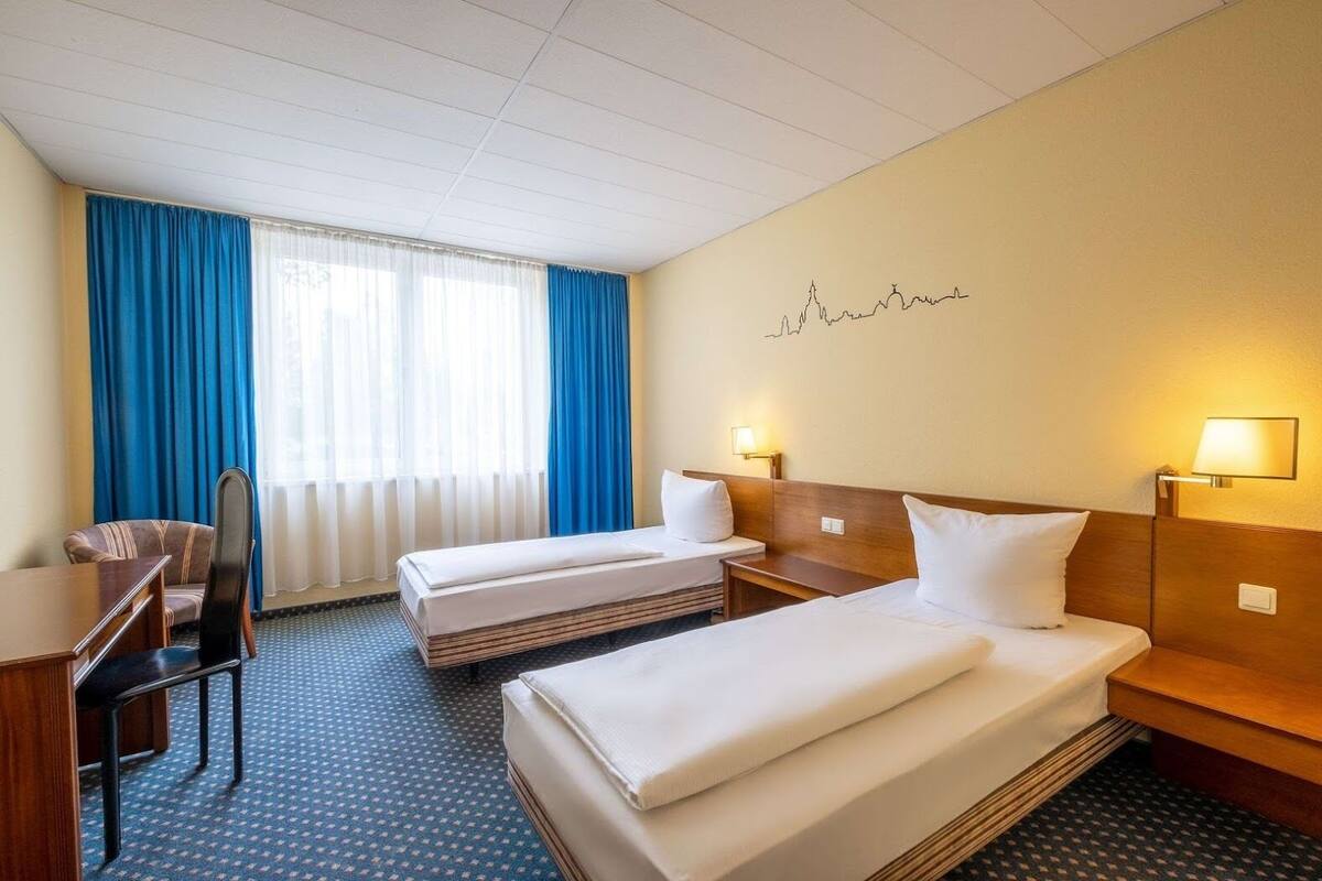 Ein Hotelzimmer mit zwei Betten, einem Schreibtisch, einem Stuhl und einem Fenster mit blauen Vorhängen.