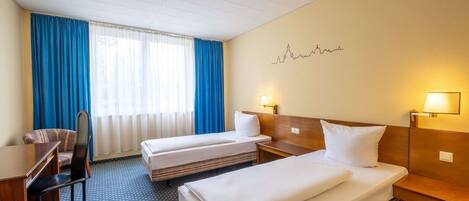 A hotel room with two beds, a desk, a chair, and a window with blue curtains.