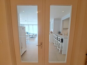 Blick durch eine verspiegelte Tür in eine moderne Küche mit weißen Schränken, einen Essbereich mit weißen Stühlen und ein sichtbares Wohnzimmer im Hintergrund.
