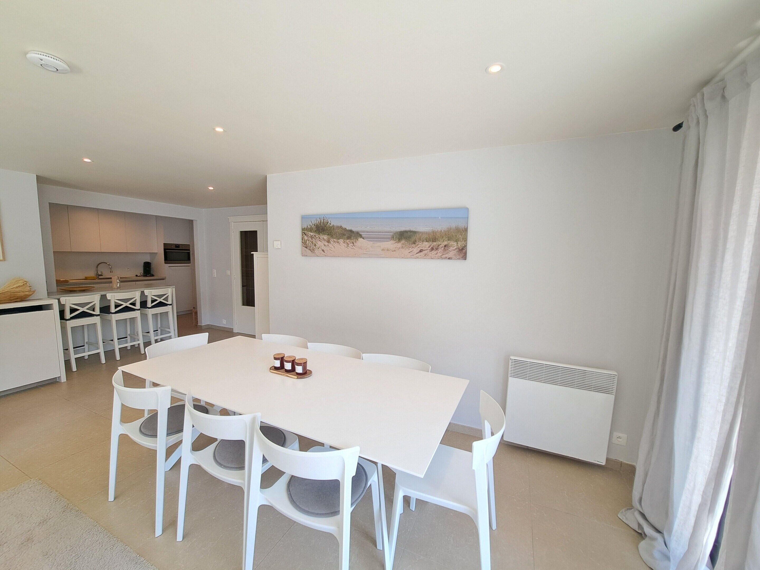 A modern kitchen with white cabinets, a dining table with four chairs, and a beach-themed painting on the wall.