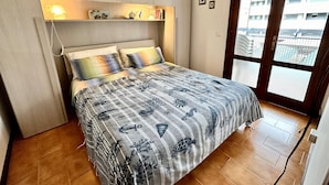 Ein Schlafzimmer mit einem Bett, einem Nachttisch, einer Ablagefläche mit Dekoration und einem Fenster mit Blick auf Gebäude, sowie einer Tür.