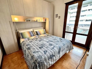 Ein Schlafzimmer mit einem Bett, einem Nachttisch, einer Ablagefläche mit Dekoration und einem Fenster mit Blick auf Gebäude, sowie einer Tür.