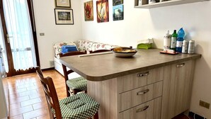 Eine kleine Küche mit Essbereich, einer Couch und einem Tisch mit Schüssel und Tellern. An der Wand befinden sich gerahmte Bilder und ein Regal mit Flaschen.