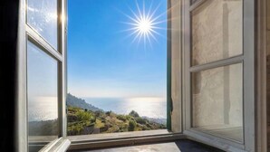 Een open raam met uitzicht op een kustlandschap en een felle zon.