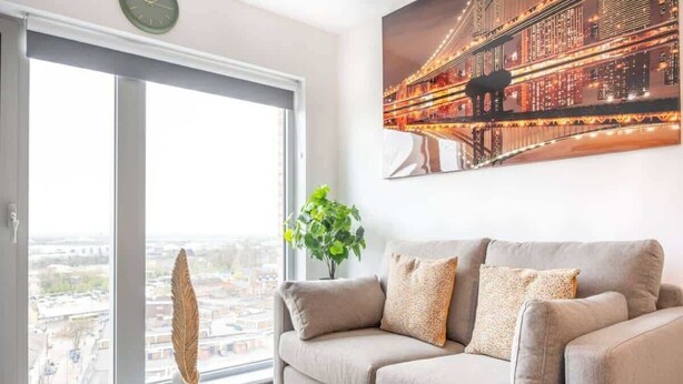 A modern living room with a sofa, a decorative feather, a plant, and a cityscape view through a large window. - Bright Apartment Near Shops in Eccles Manchester (Eccles)