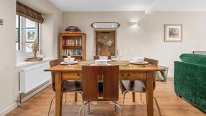 Un coin repas avec une table en bois dressée pour quatre personnes, une bibliothèque, une fenêtre donnant sur un beau paysage et, au fond, un canapé vert.