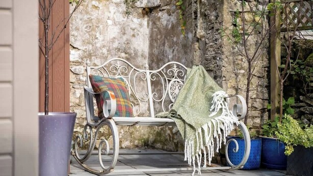 Un banc en métal agrémenté d’un coussin coloré, d’un plaid vert et de plantes en pot.
