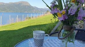 A table with a glass mug and a vase of flowers, overlooking a lake with mountains in the background. - Serene Fjord View Holiday House in Davik, Nordfjord (Bremanger)