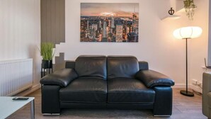 Ein modernes Wohnzimmer mit einer schwarzen Ledersofa, einem Stadtbild an der Wand, einer Stehlampe und einem Topf mit Pflanze.