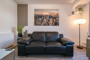 Un salón moderno con un sofá de cuero negro, un cuadro de un paisaje urbano en la pared, una lámpara de pie y una planta en maceta.