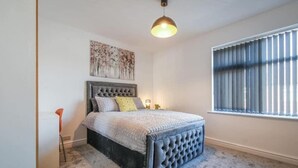 Ein Schlafzimmer mit einem gepolsterten Kopfteil, einem Bett mit grauer Bettdecke, einem Nachttisch, einer Stehlampe und einem Fenster mit Jalousien.
