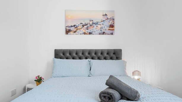 Un letto rifatto con un comodo poggiatesta grigio, un asciugamano arrotolato e un quadro che raffigura una cittadina costiera.