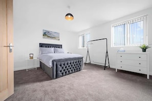 Un dormitorio con cama, mesita de noche, lámpara, cómoda, ventana y un cuadro en la pared.