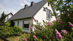 Een wit huis met een donker dak, omringd door groen en bloeiende planten.