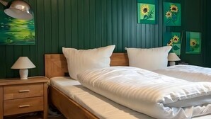 Een tweepersoonsbed met witte beddengoed, een houten hoofdeinde, een nachtkastje met lamp, en een groene muur met zonnebloemkunstwerk.