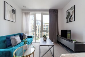 Ein modernes Wohnzimmer mit einem türkisfarbenen Sofa, einem Glastisch, einem Flachbildfernseher auf einem schwarzen Standfuß und einem großen Fenster mit Vorhängen.
