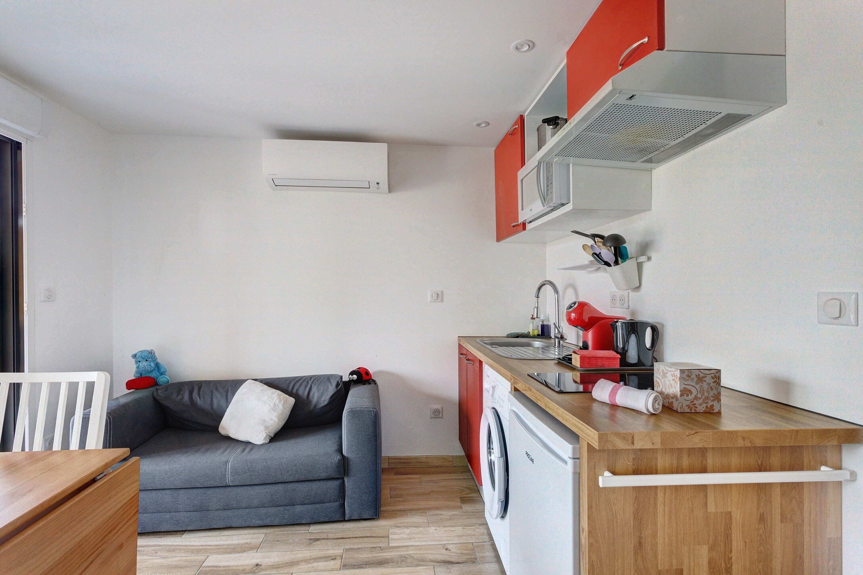 Un espace de vie compact comprenant un canapé gris, un lave-linge, une table en bois et un plan de travail de cuisine rouge.