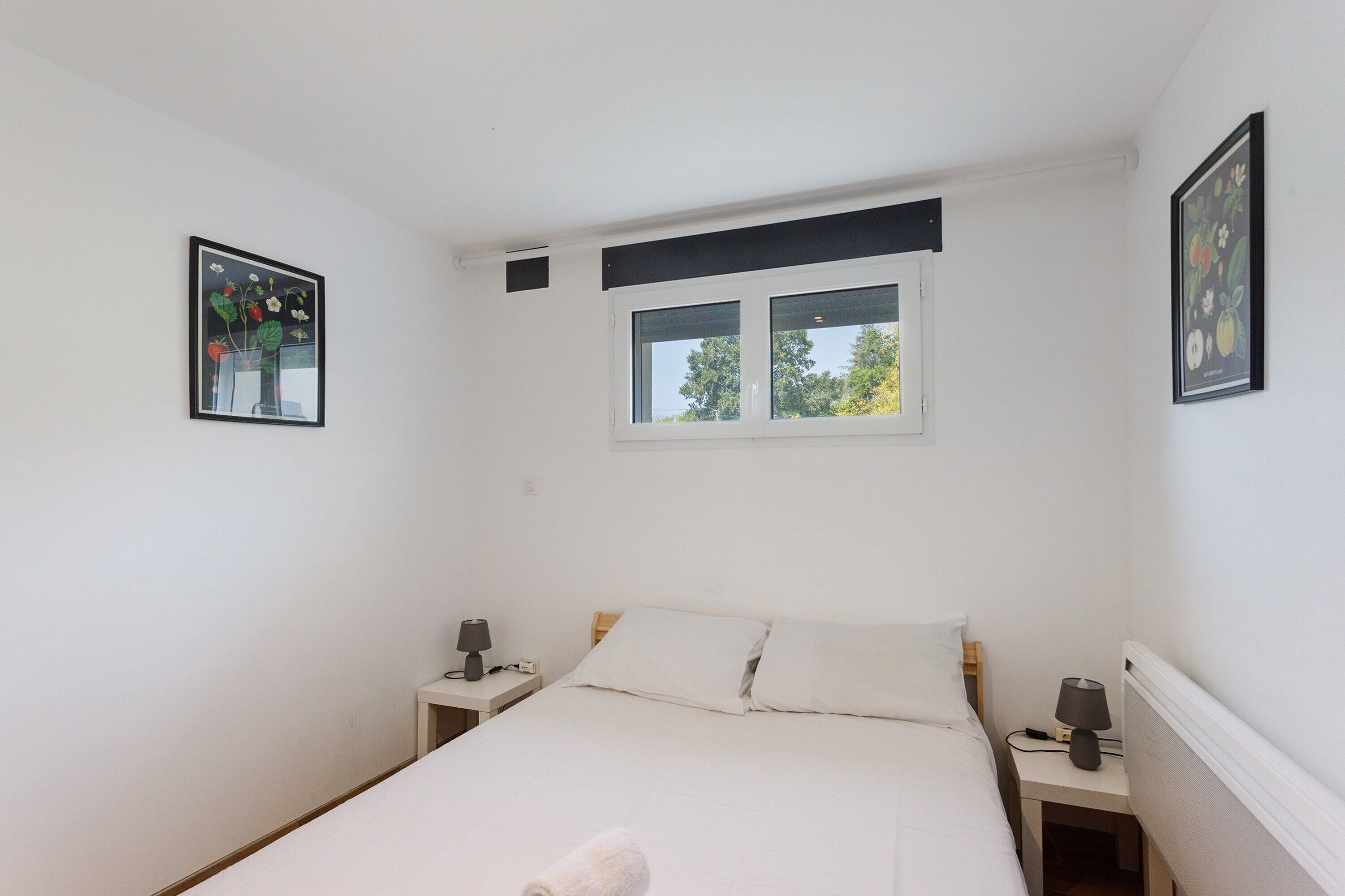 A single bed with white linens, two bedside tables with lamps, a window with a view of trees, and two framed artworks on the walls.