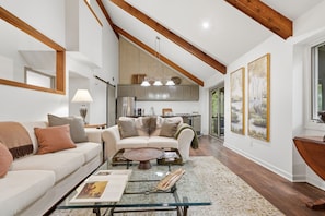 A spacious living room with a glass coffee table, a sofa with cushions, a kitchen in the background, and artwork on the walls. - Lakefront Tree House | Big Canoe Mountain Getaway (Jasper)