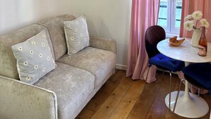 Ein gemütliches Wohnzimmer mit einer Couch, einem Tisch mit einer Vase und einer Flasche, und Blick durch rosa Vorhänge.