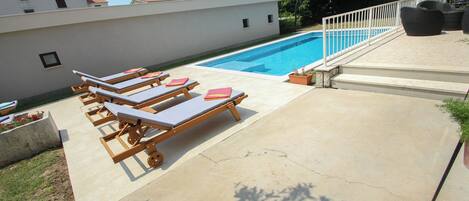 Una piscina con sillones de descanso, una cerca blanca y un edificio al fondo.